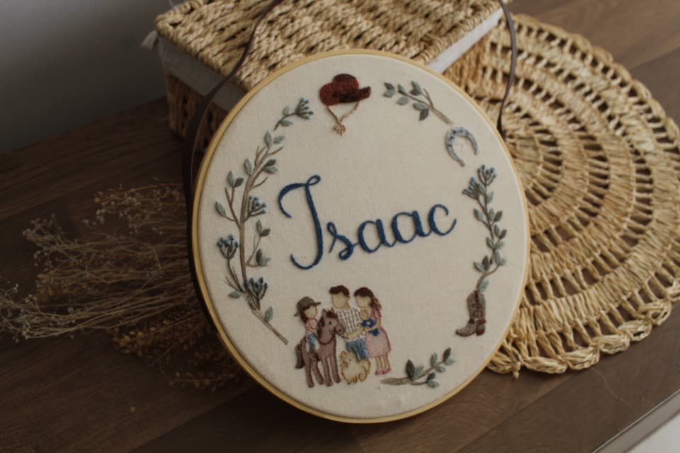 Porta-maternidade em bastidor com o nome Isaac, guirlanda de folhas e família bordada